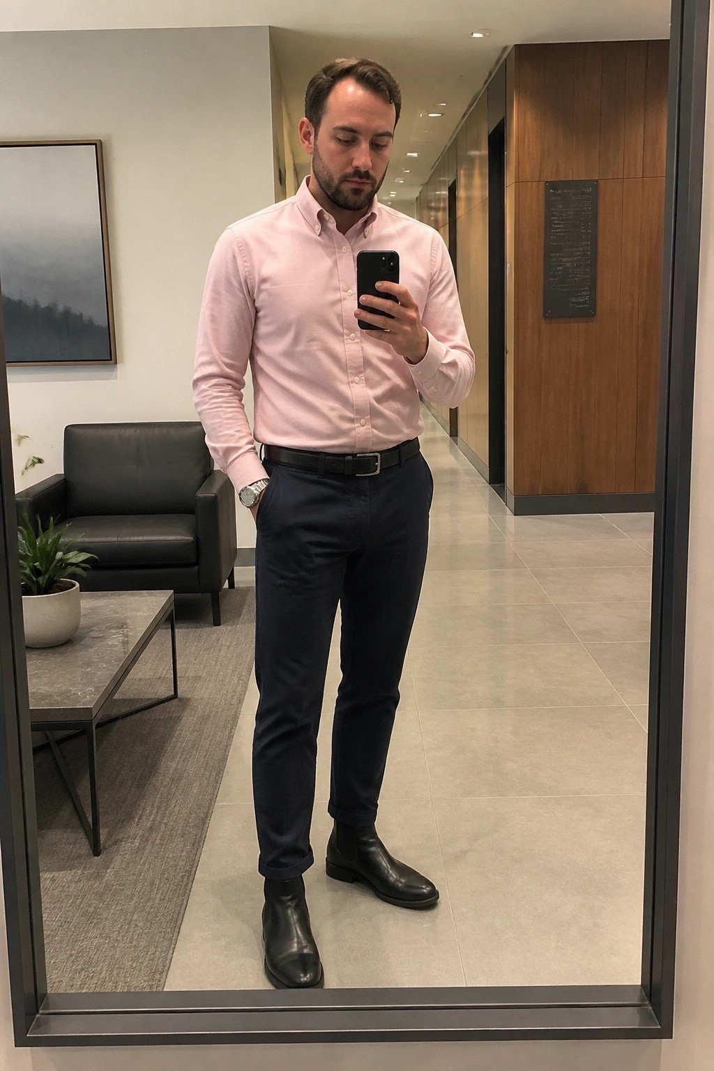 Man in pink shirt and navy pants taking mirror selfie in office hallway.