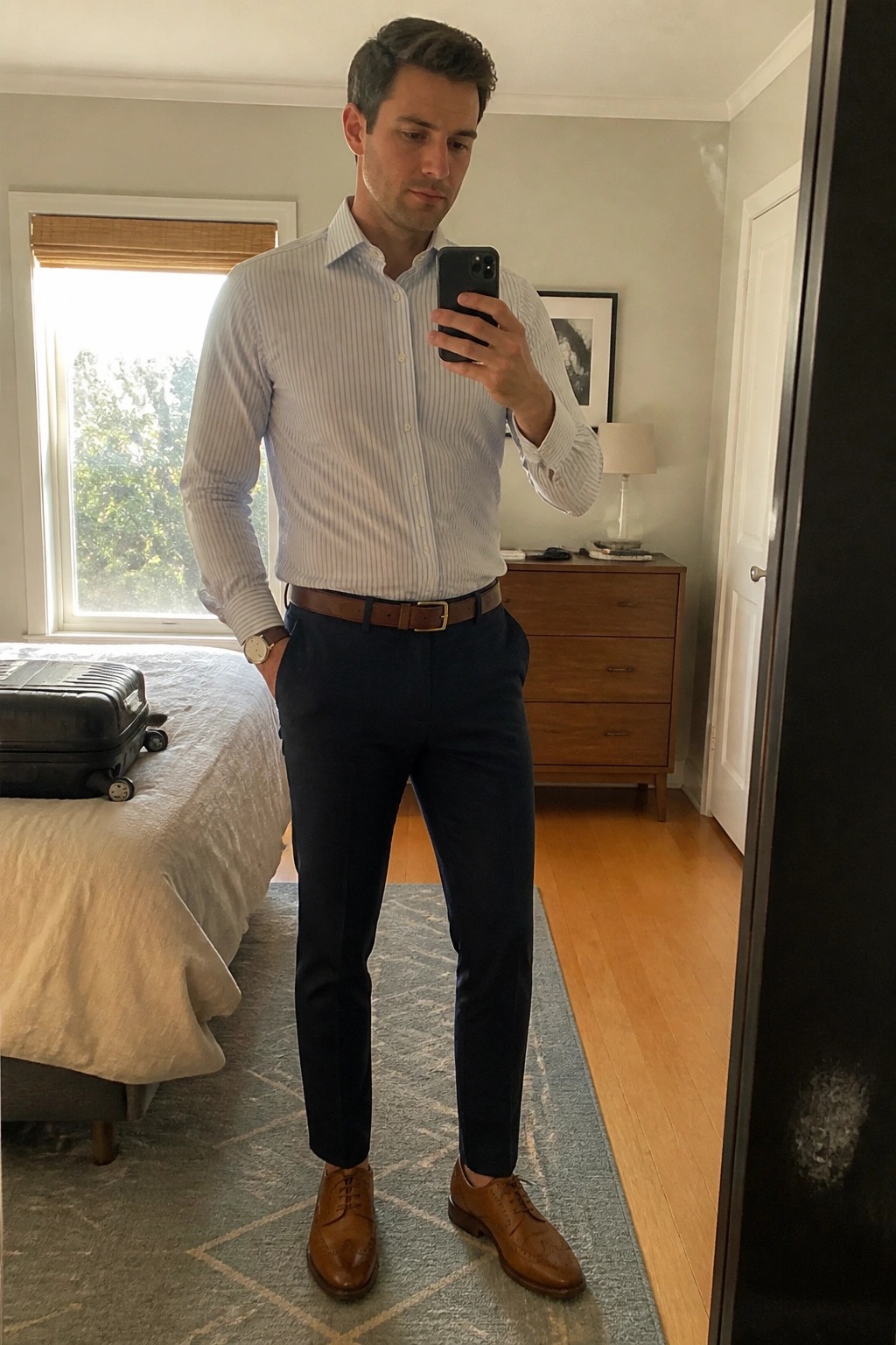 Man in light blue striped shirt and navy pants taking mirror selfie in bedroom