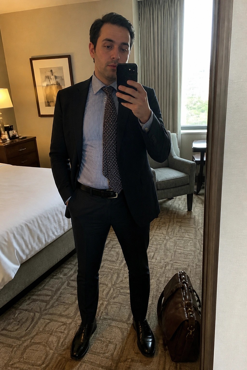Man in navy suit and tie taking mirror selfie in hotel room with leather bag