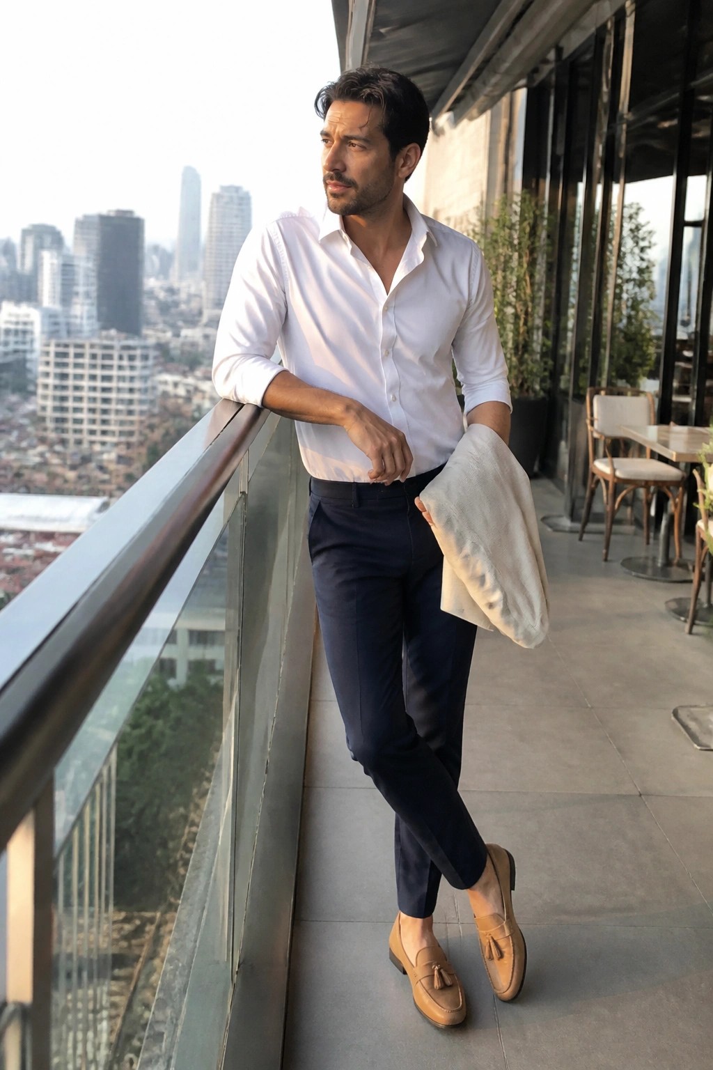 Man in white shirt and navy pants leaning on balcony overlooking cityscape