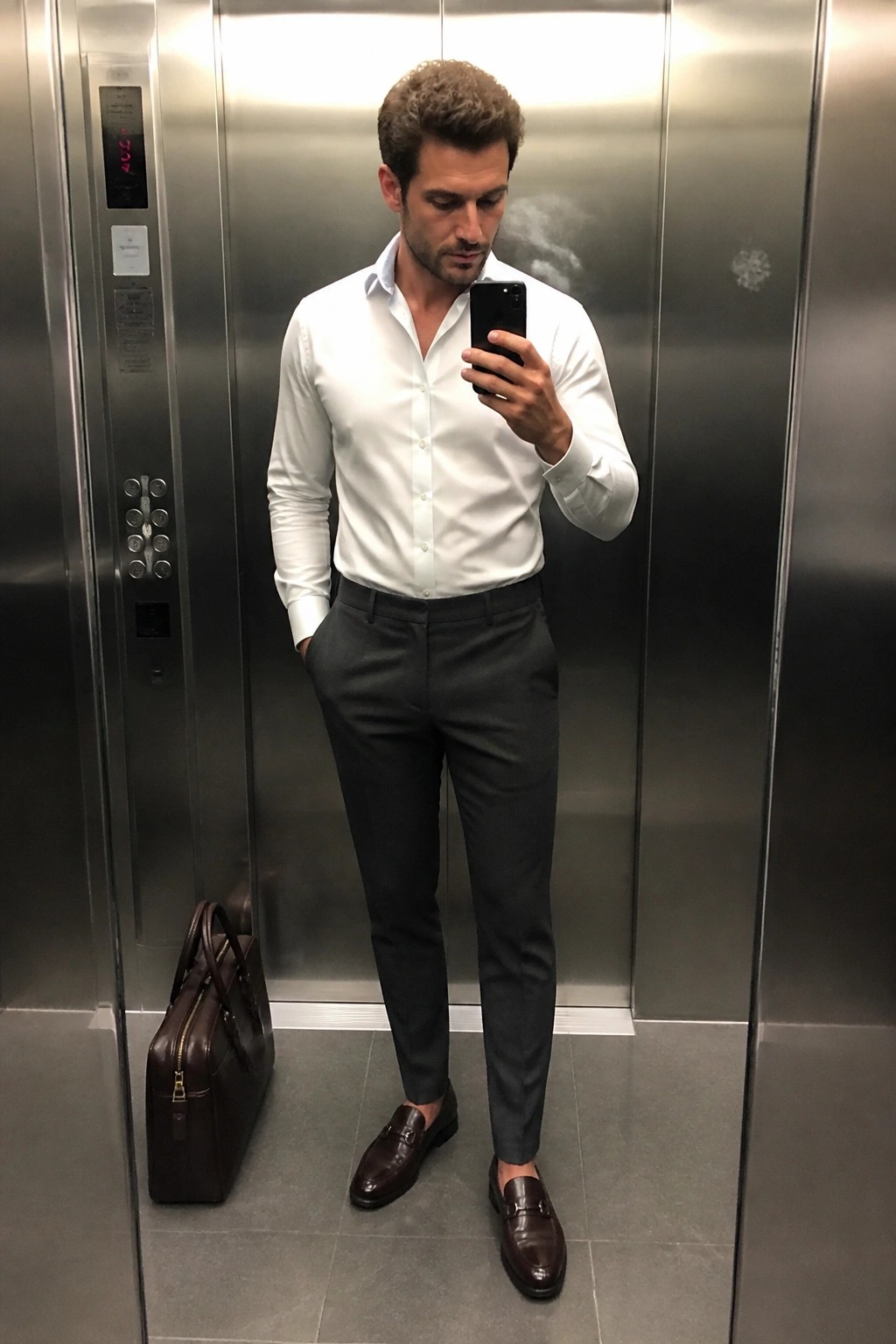 Man in white shirt and gray pants taking selfie in elevator with briefcase.