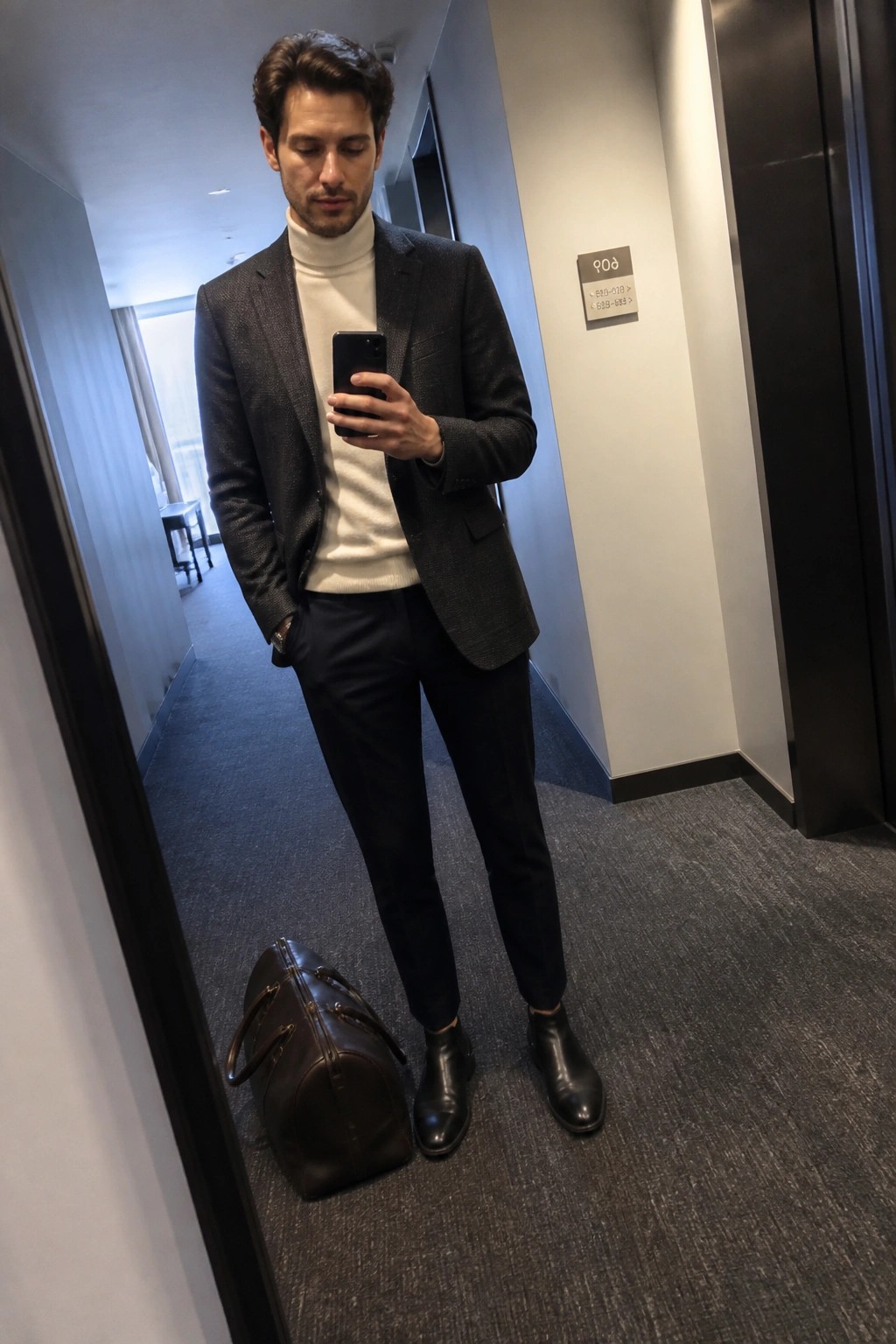 Man in dark blazer over cream turtleneck checks phone beside leather bag in hallway