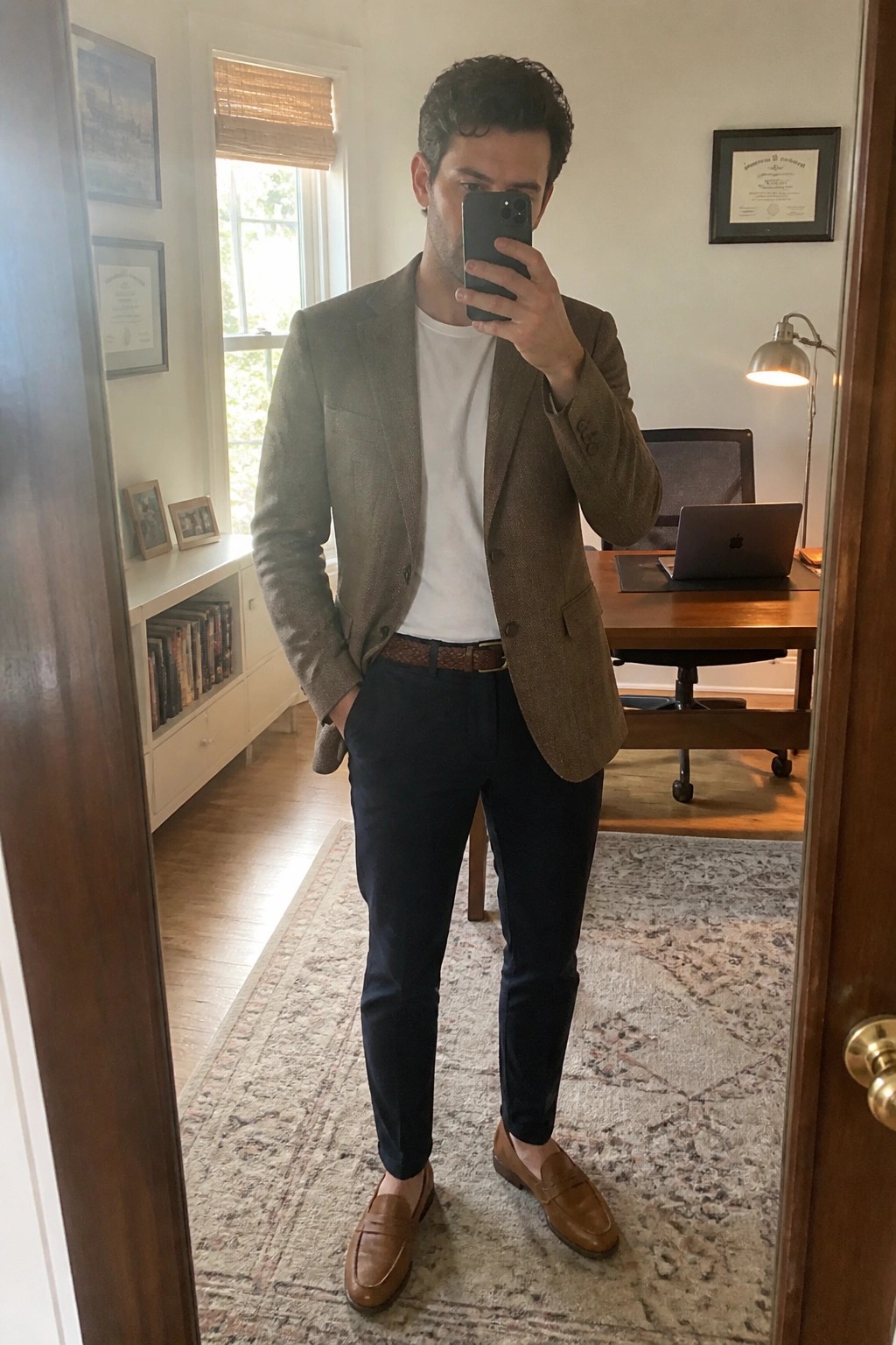 Man in brown tweed blazer, white tee, navy pants takes mirror selfie in office