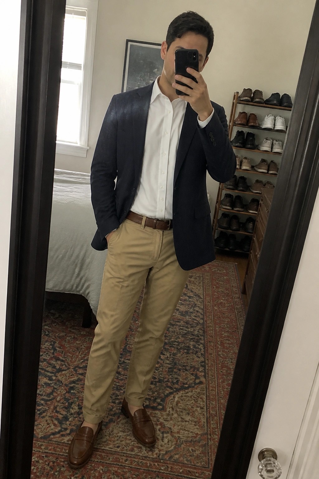 Man in navy blazer, white shirt, and khaki pants taking bedroom mirror selfie.