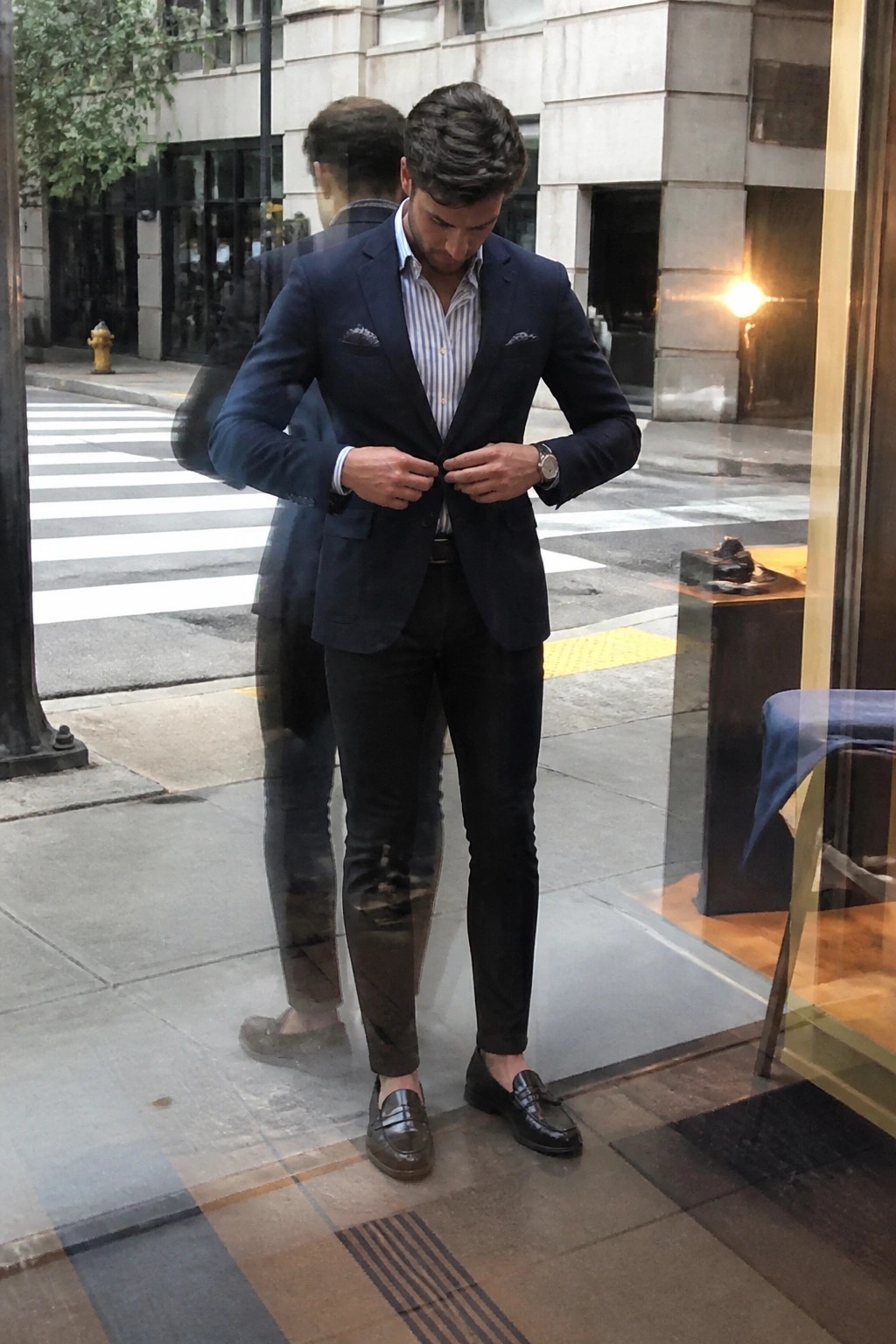 Man in navy suit adjusting jacket buttons on city sidewalk with window reflection