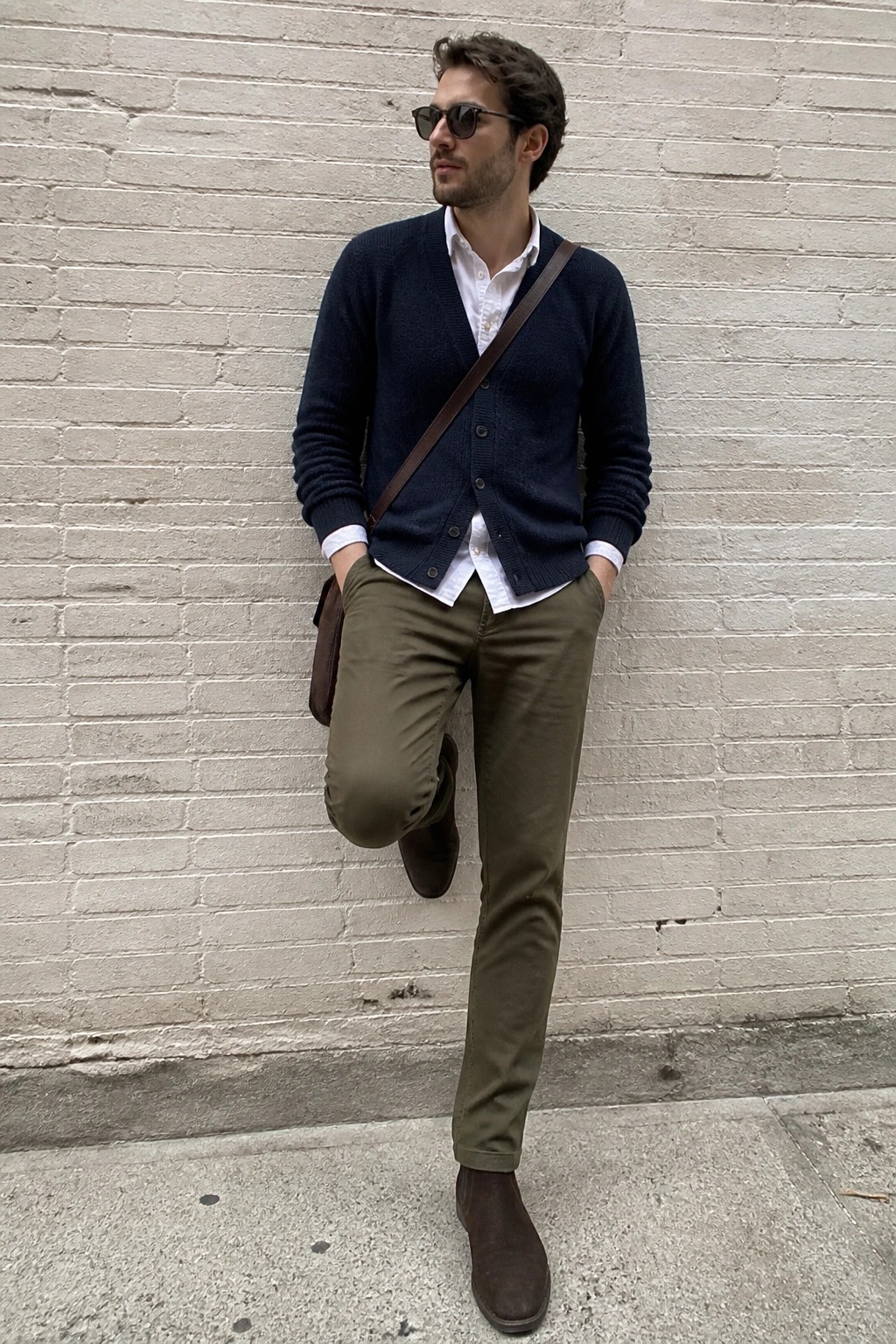 Man in navy cardigan, white shirt, olive pants, and brown boots by brick wall with bag.