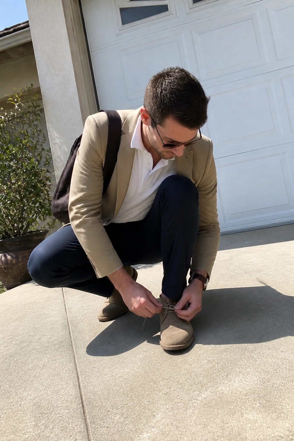Man in tan blazer squats tying laces on suede boots by white garage