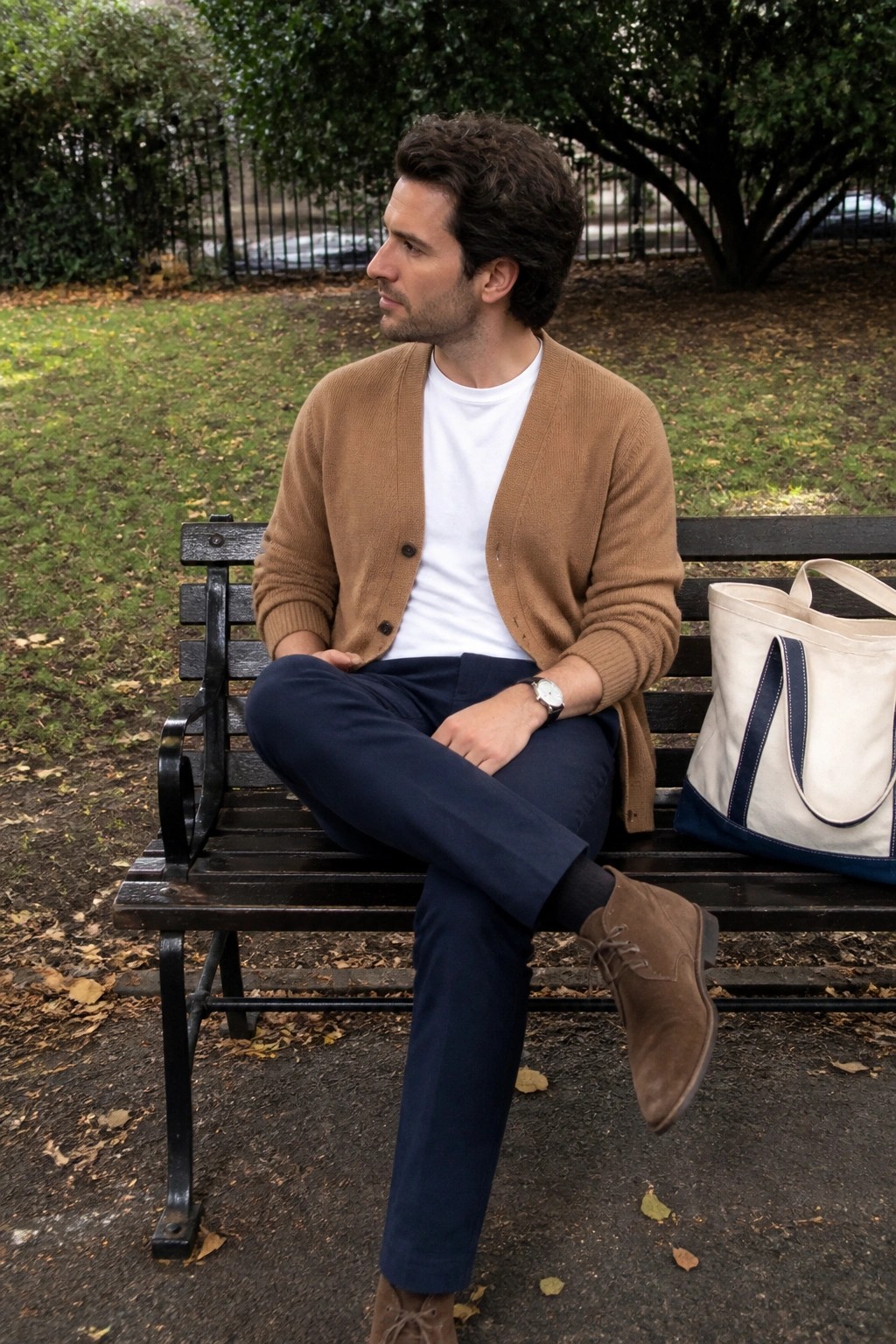 Man seated on park bench wearing open brown cardigan over white t-shirt, navy trousers, brown suede boots, and wristwatch with canvas tote beside him.