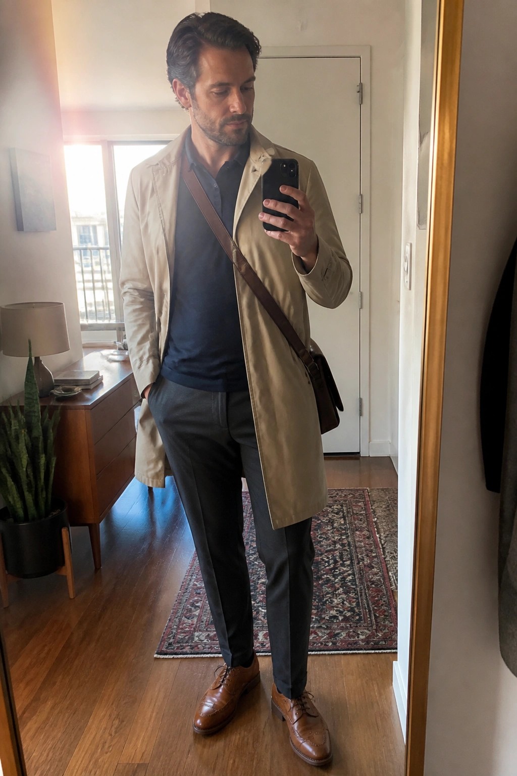 Man in beige trench coat over navy shirt and gray pants taking mirror selfie