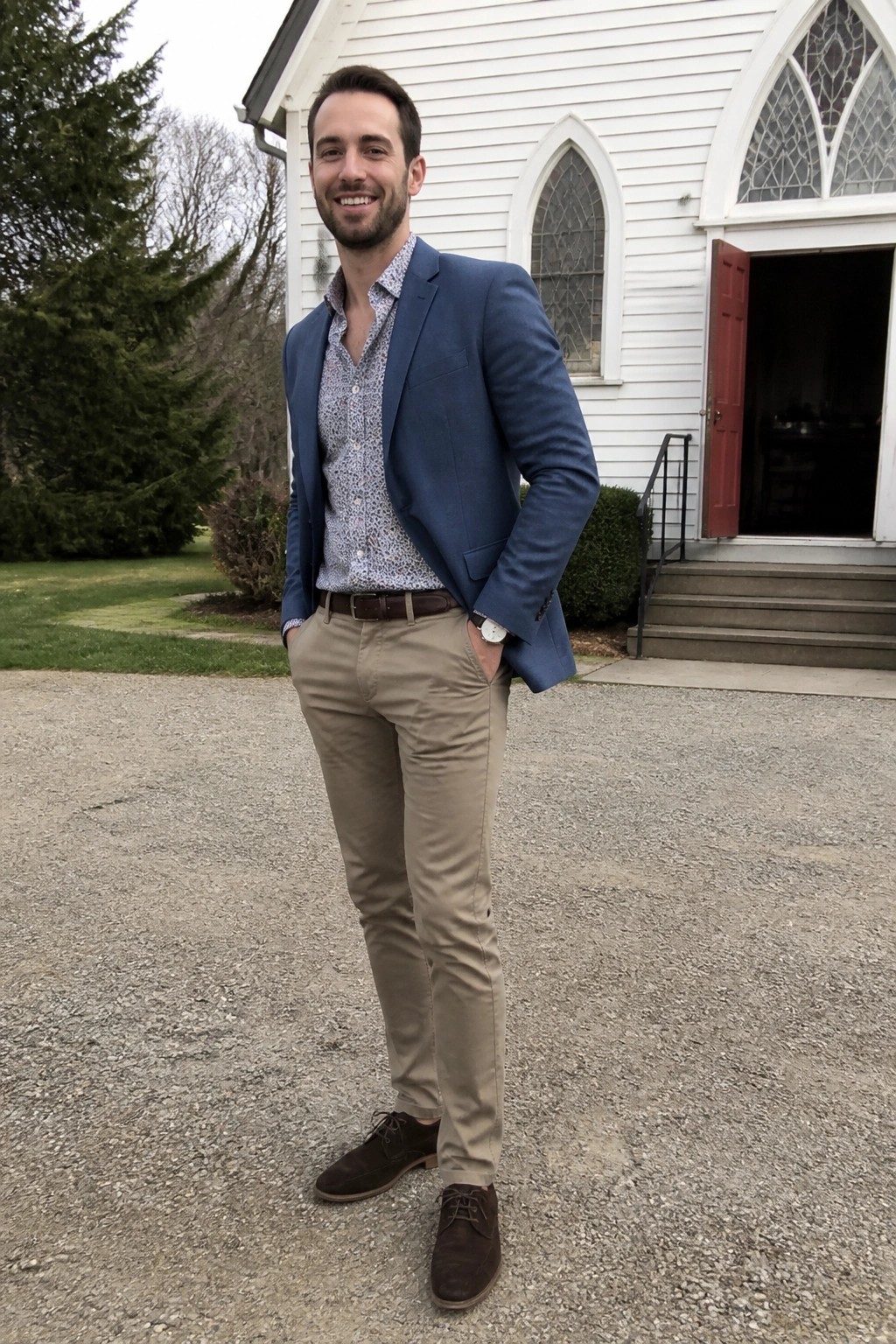 Smiling man in blue blazer and khakis stands before white church with red door.