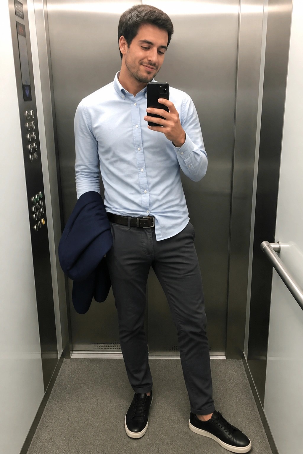 Man in light blue shirt and gray pants taking selfie in elevator holding navy jacket
