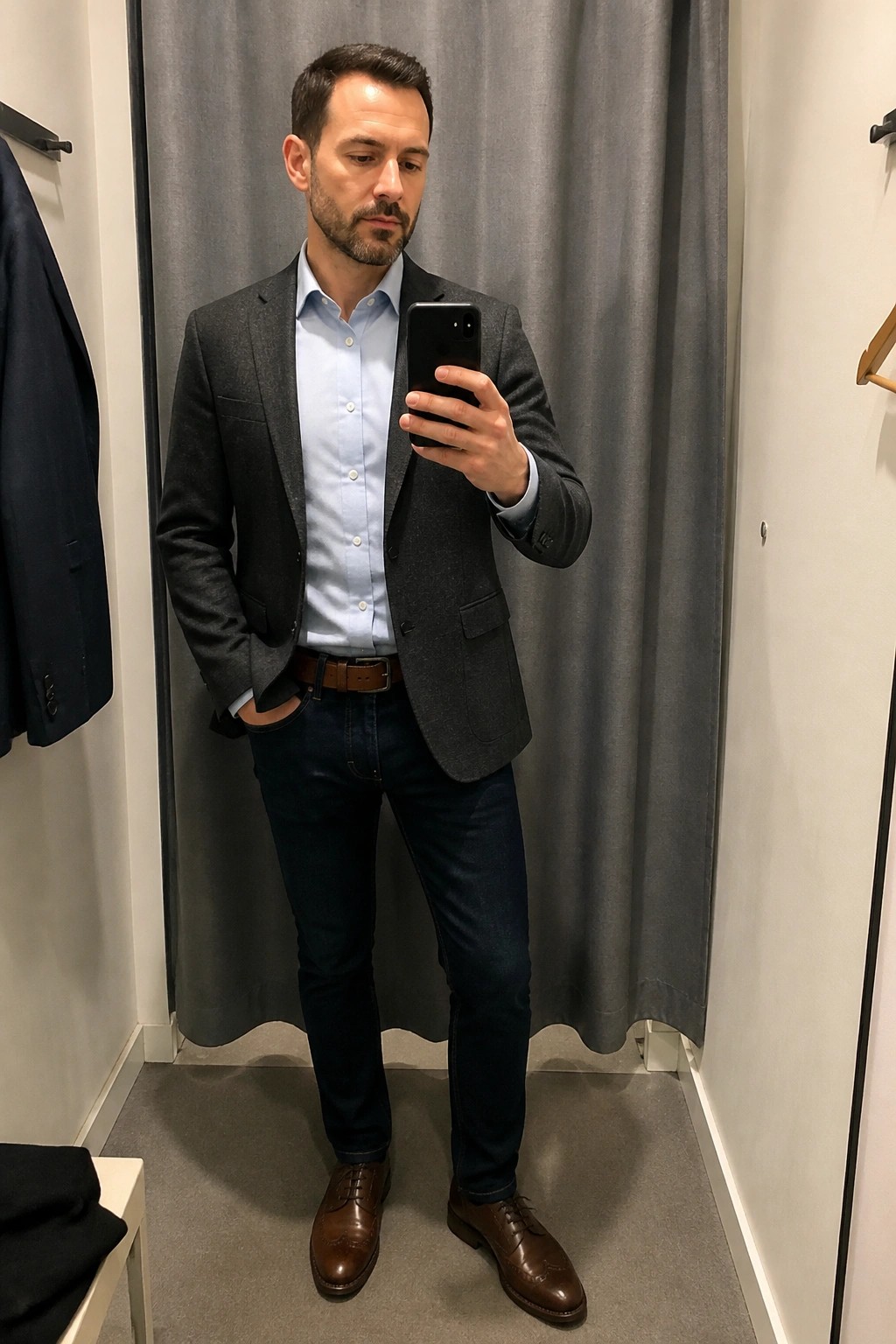 Man in gray blazer and light blue shirt taking mirror selfie in fitting room