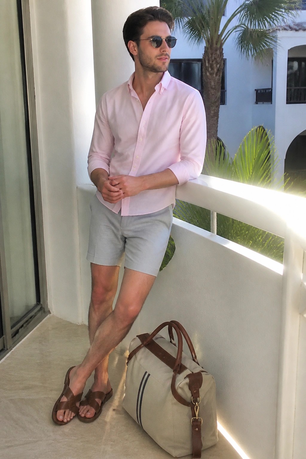 Man in pink shirt and gray shorts leaning on balcony beside duffel bag