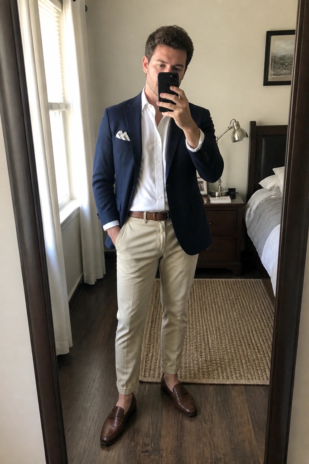 Man in navy blazer, white shirt, and beige pants taking mirror selfie in bedroom
