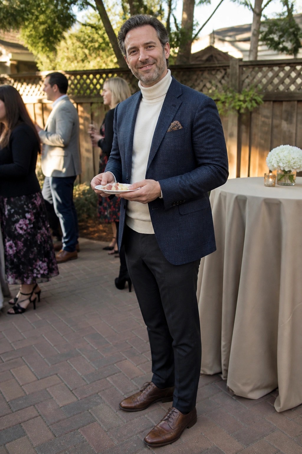 Man in navy blazer and cream turtleneck holding plate at outdoor gathering