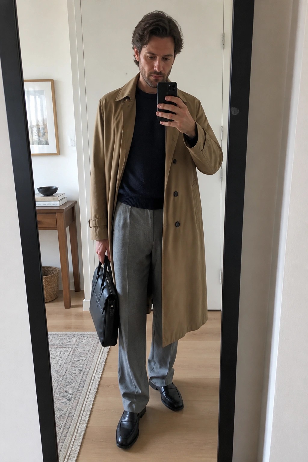 Man in tan trench coat and gray trousers takes mirror selfie with phone and bag
