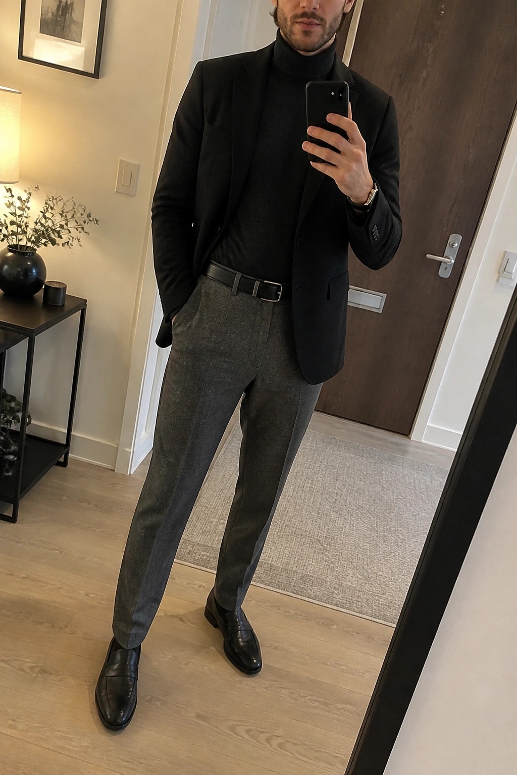 Man in black blazer over turtleneck with grey trousers taking mirror selfie.