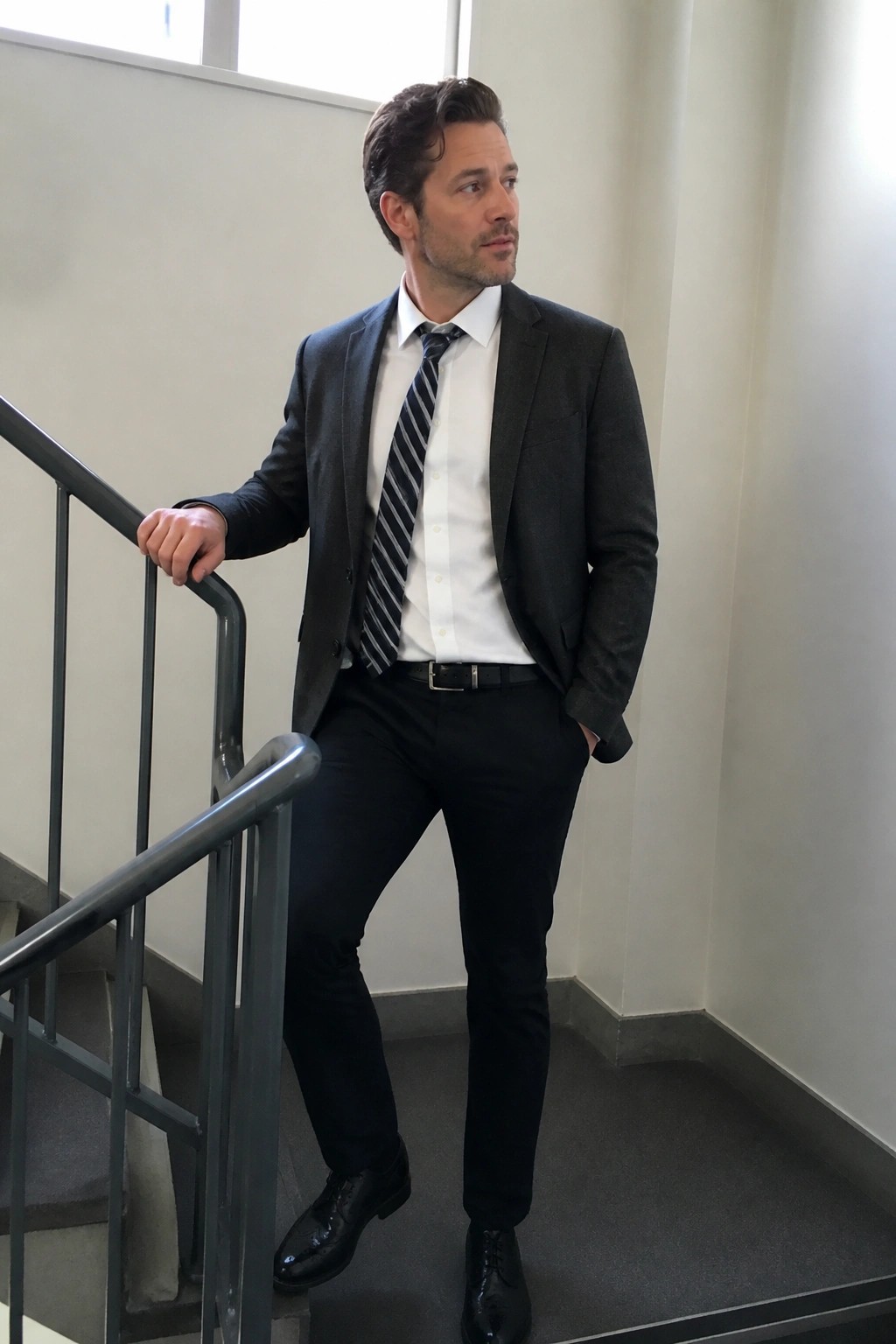 Man in dark suit and striped tie stands on stairs holding railing