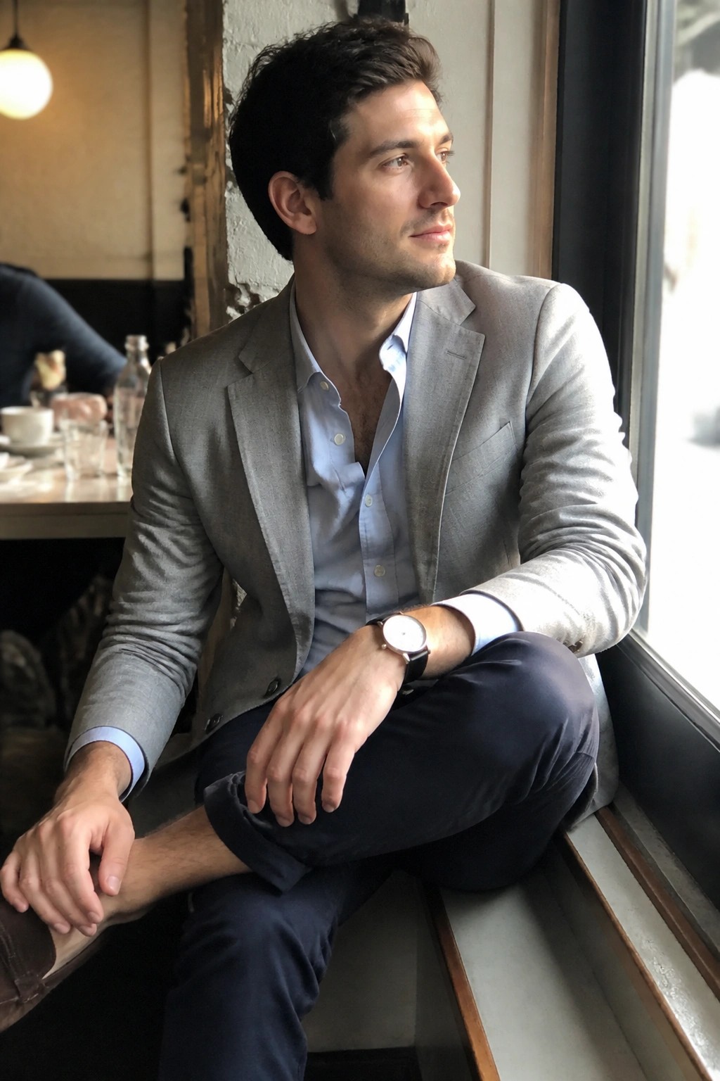 Man in gray blazer and blue shirt seated by cafe window wearing watch