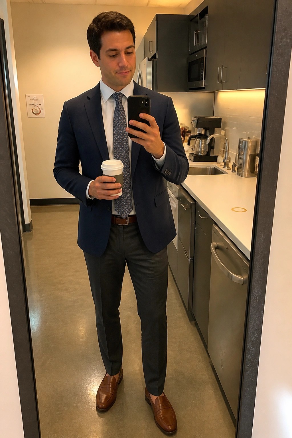 Man in navy suit holding coffee and phone taking mirror selfie in office kitchen