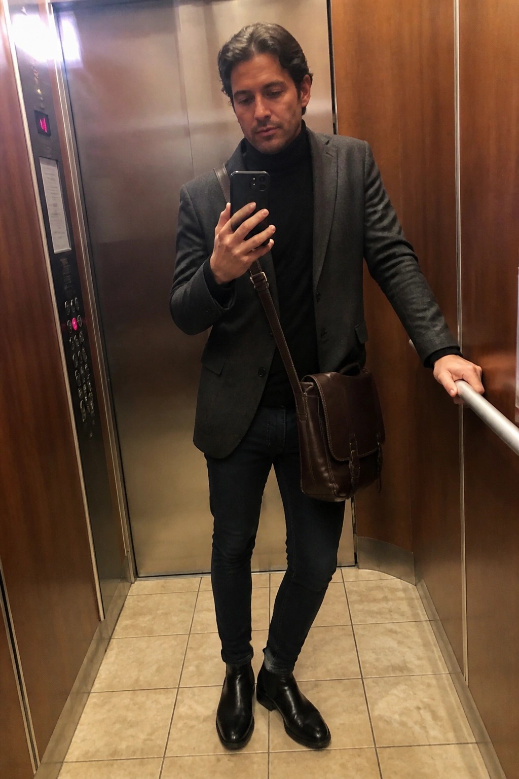 Man in gray blazer over black turtleneck holding phone in elevator with leather bag.