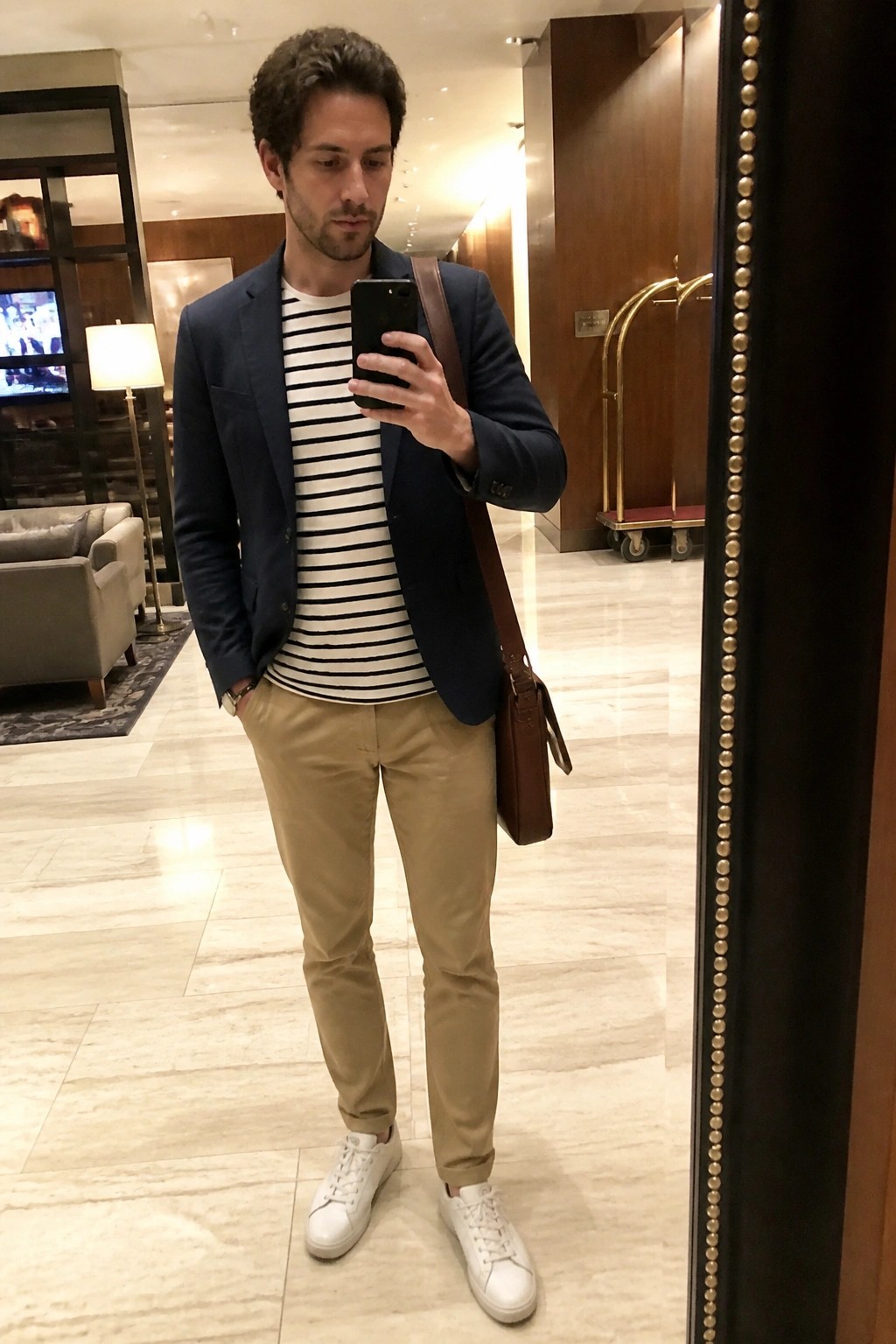 Man in navy blazer over striped tee with khakis taking mirror selfie in lobby