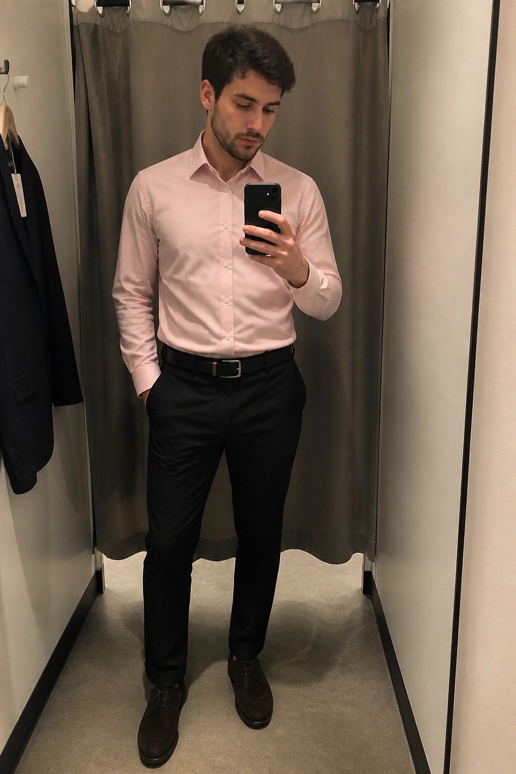 Man in light pink shirt and black pants taking mirror selfie in fitting room