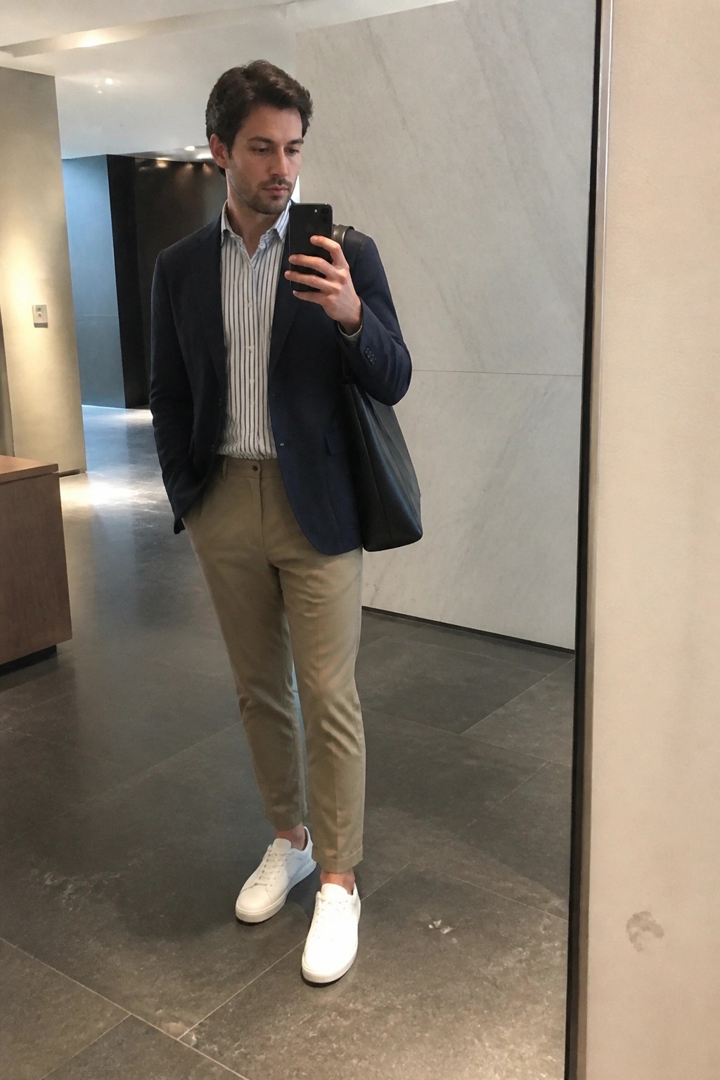 Man in navy blazer, striped shirt, beige pants, and white sneakers taking mirror selfie.