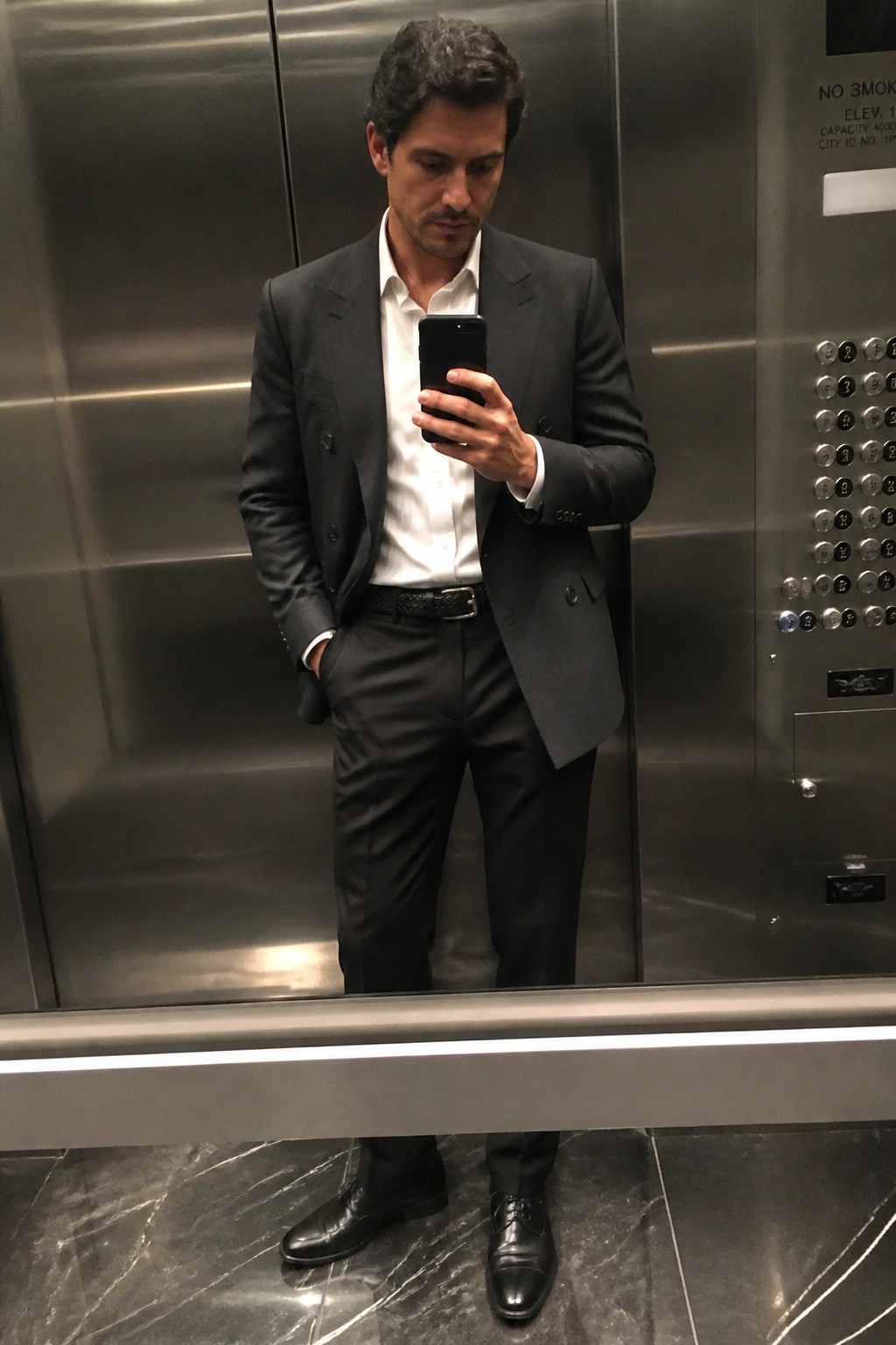 Man in dark gray suit taking mirror selfie with phone in elevator.