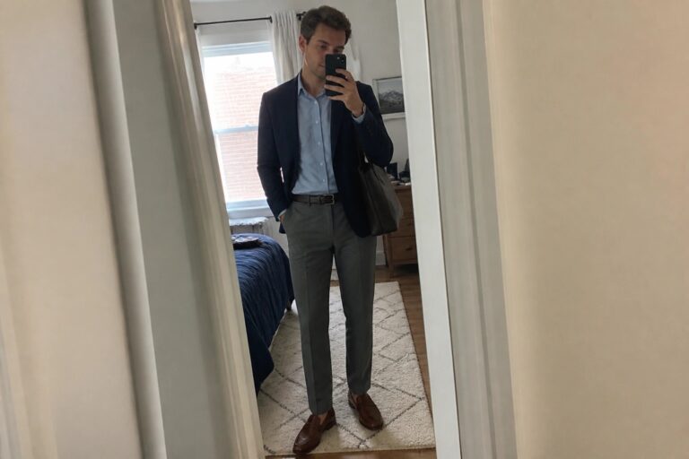 21 Elevated Men’s Office Outfits Built Around Modern Workwear Essentials Man wearing a navy blazer over a light blue button down shirt with grey trousers and brown loafers while holding a black bag in a bedroom mirror selfie.