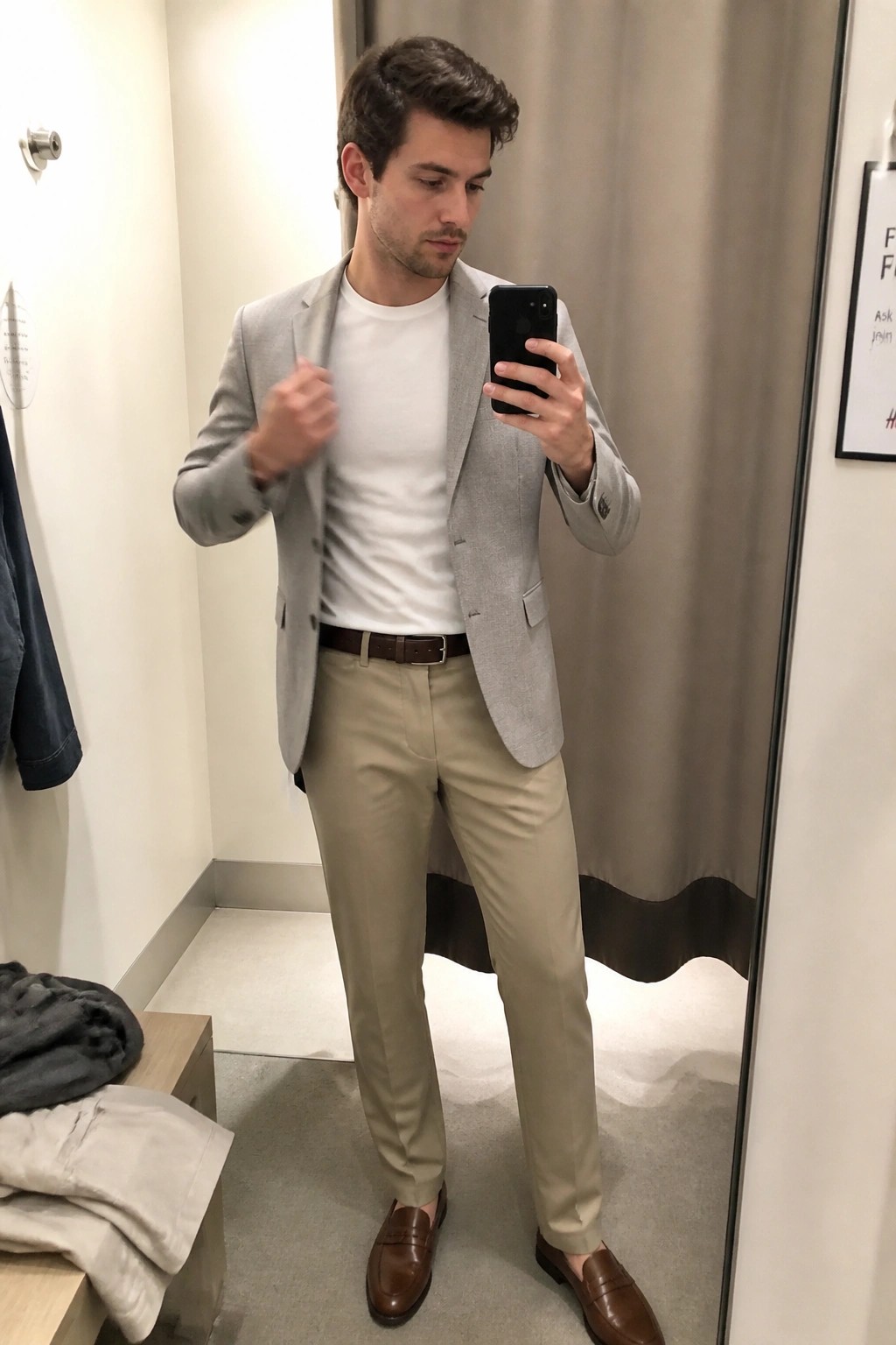 Man in gray blazer, white t-shirt, khaki pants taking mirror selfie in fitting room