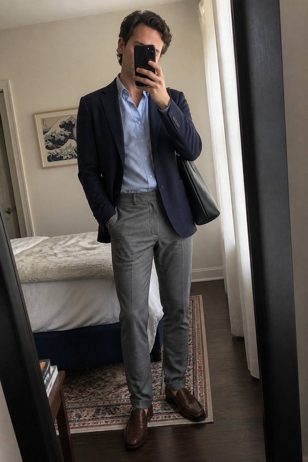 Man wearing a navy blazer over a light blue button down shirt with grey trousers and brown loafers while holding a black bag in a bedroom mirror selfie.