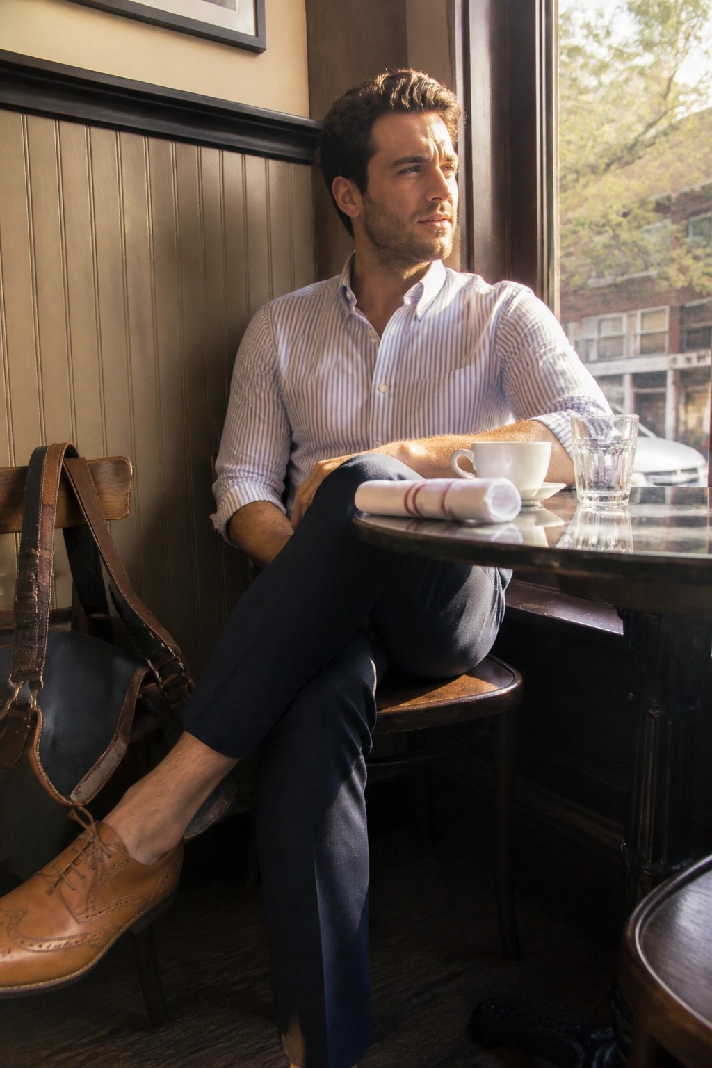 Man in striped shirt seated at cafe table by window with coffee and bag