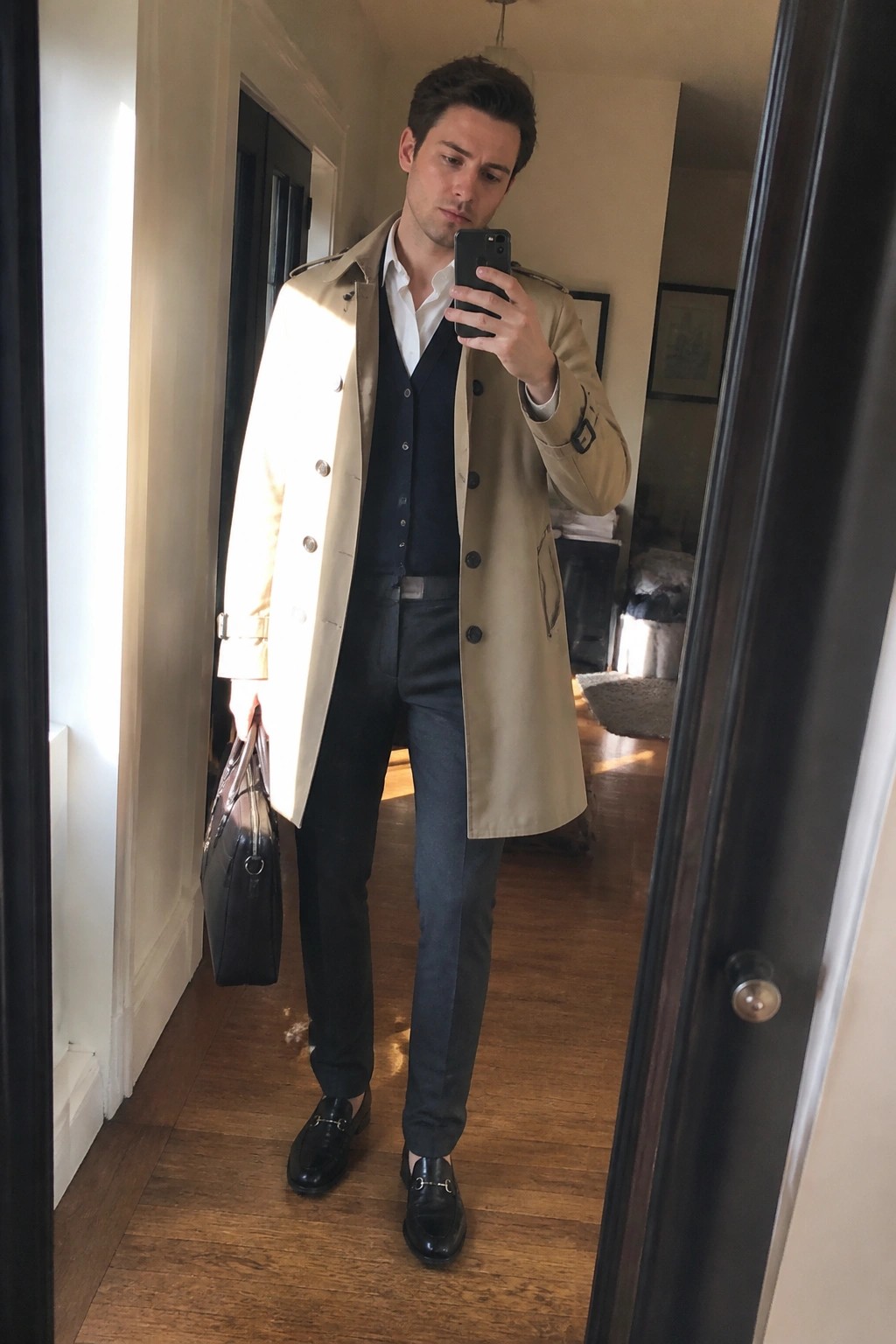 Man in beige trench coat and navy vest holding briefcase during mirror selfie in hallway.