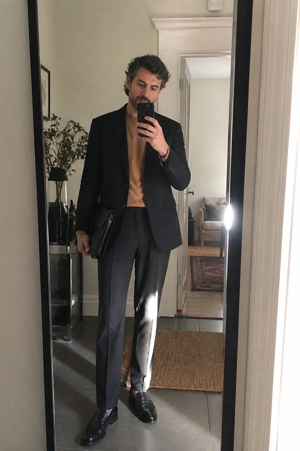 Man in black blazer and tan sweater taking mirror selfie with phone and clutch