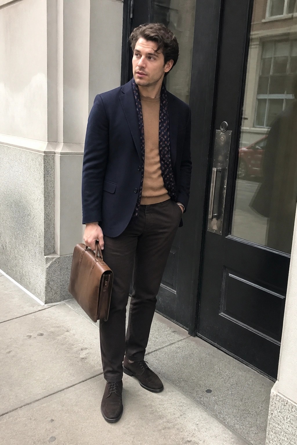 Man in navy blazer and tan sweater holding brown briefcase outside black door.