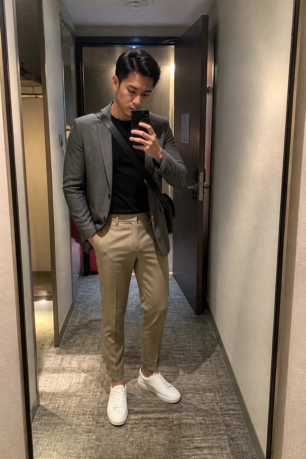 A man in a gray blazer over black tee with beige trousers and white sneakers taking a mirror selfie in a hallway.