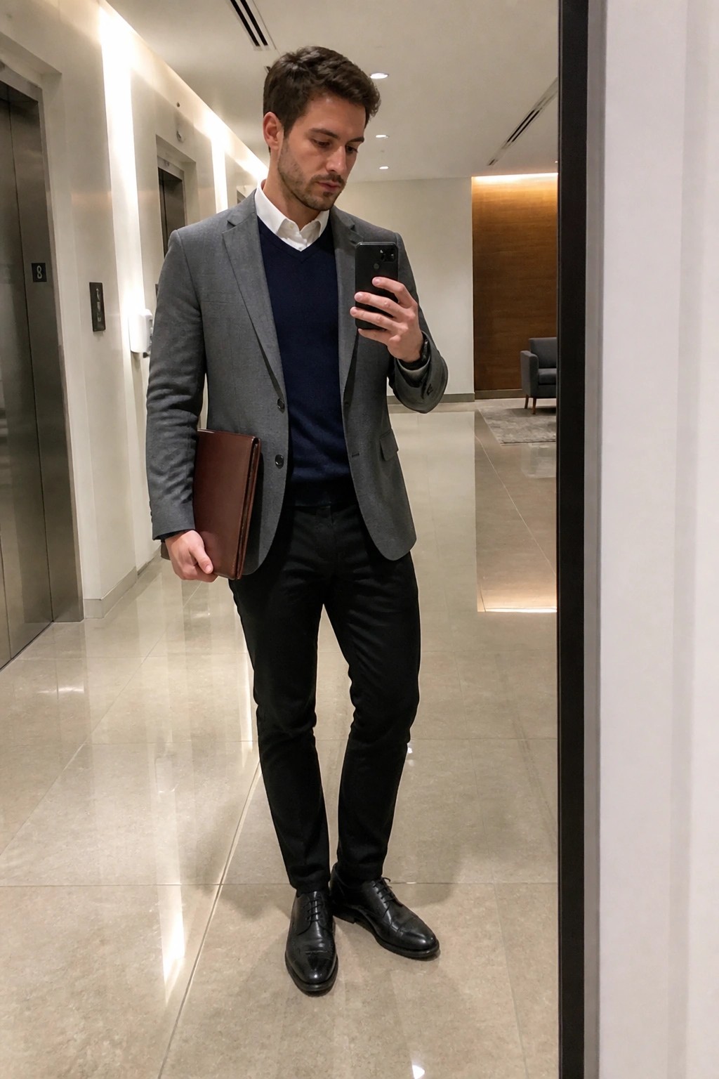 Man in gray blazer over navy sweater holding smartphone and brown portfolio in hallway.