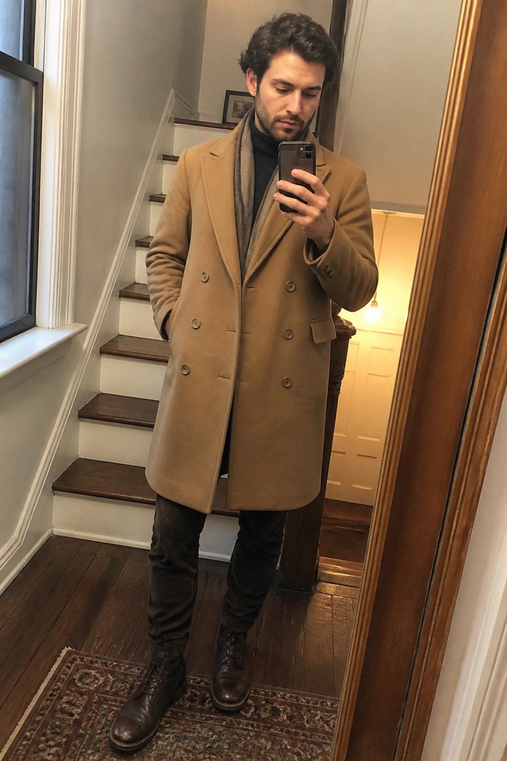Man in tan double-breasted overcoat and scarf taking mirror selfie with phone