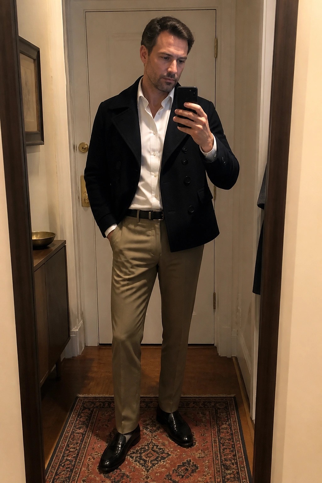 Man in black double-breasted jacket, white shirt, khaki pants takes mirror selfie.