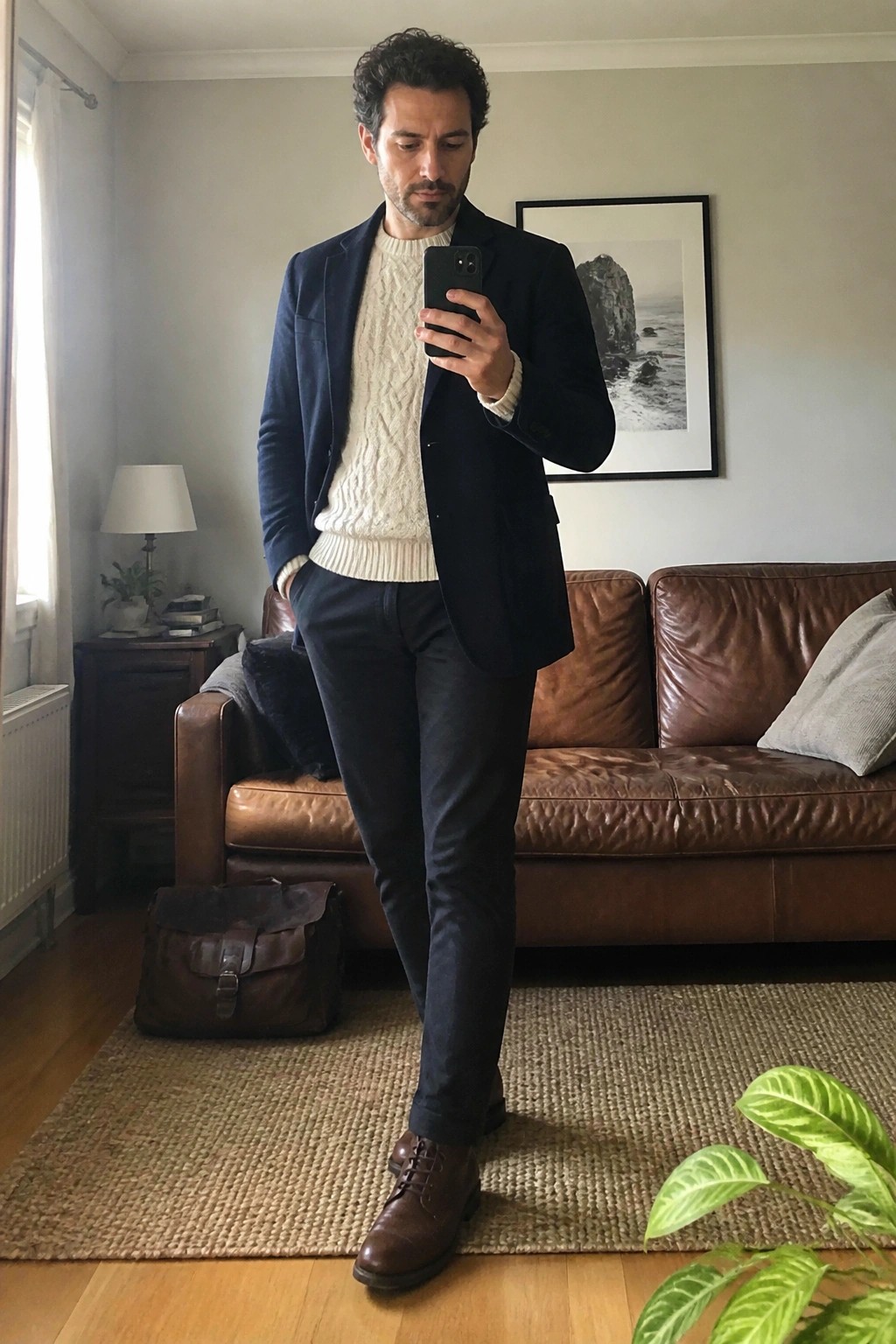 Man in navy blazer over cream cable-knit sweater taking mirror selfie in living room