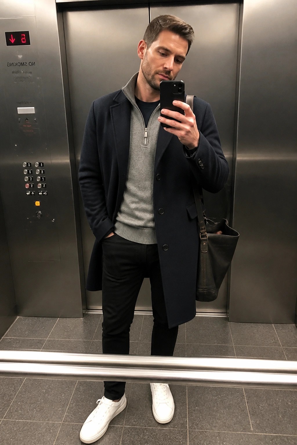 Man in navy coat over gray sweater taking mirror selfie in elevator.