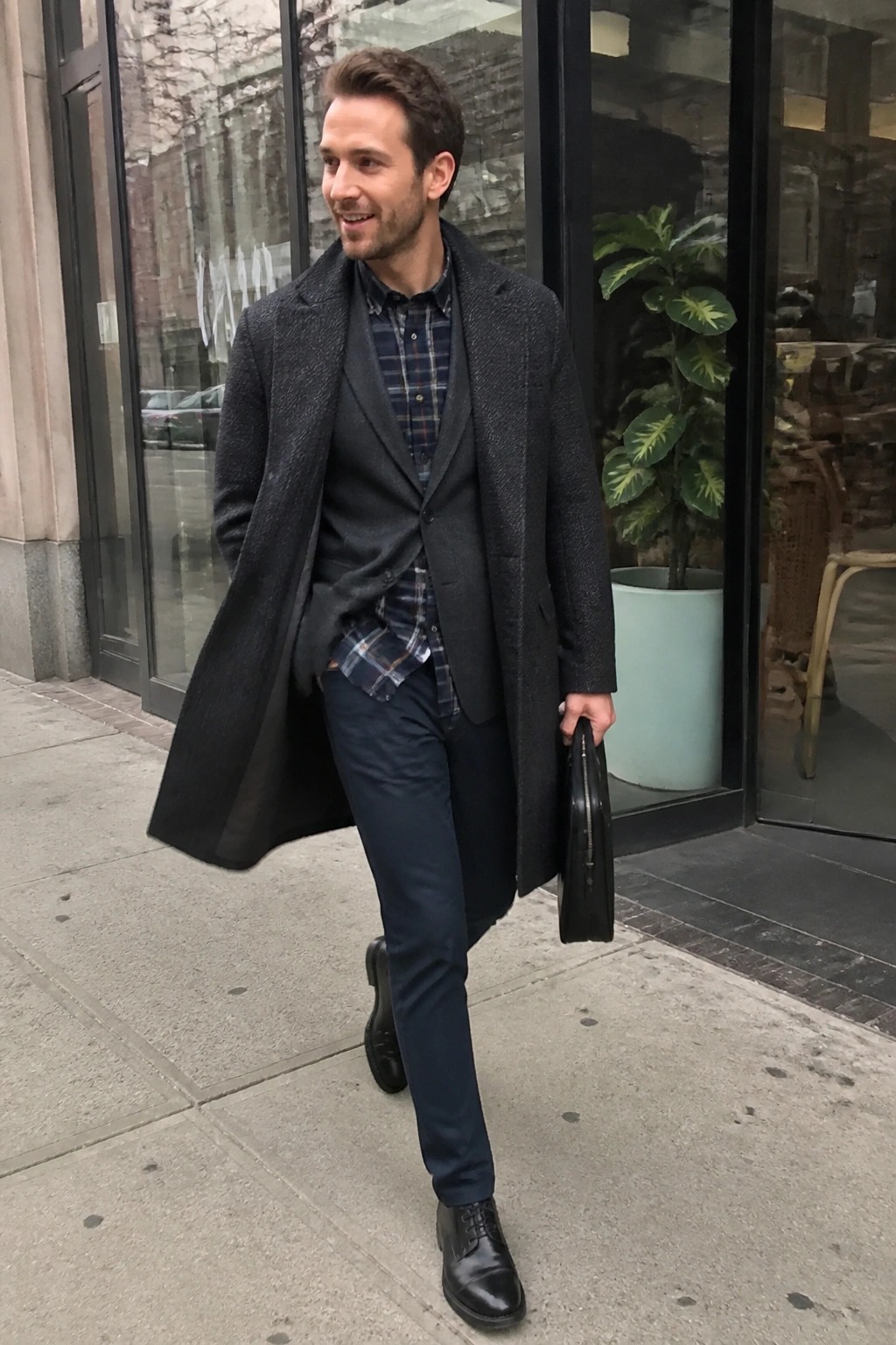 Man in dark overcoat over vest and plaid shirt carries briefcase on sidewalk