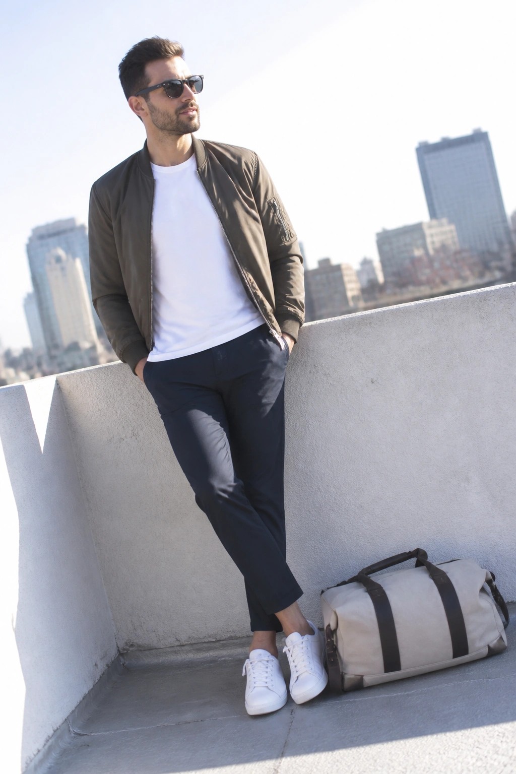 Man in green bomber jacket and white sneakers leaning on ledge with duffel bag