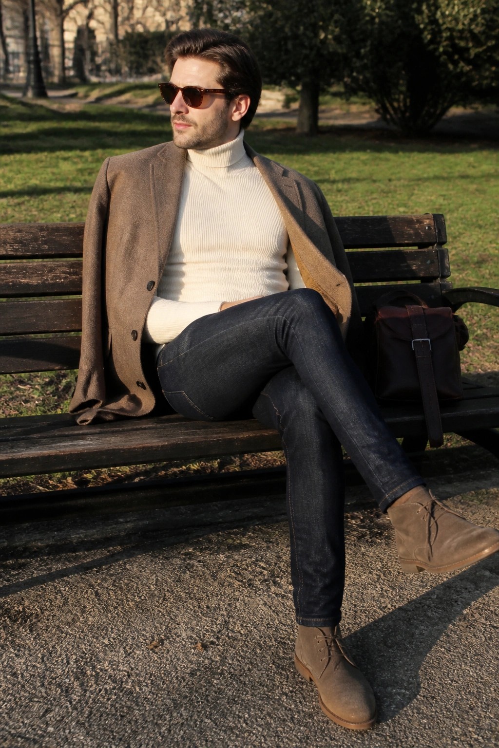 Man in brown coat over white turtleneck sits on park bench with leather bag