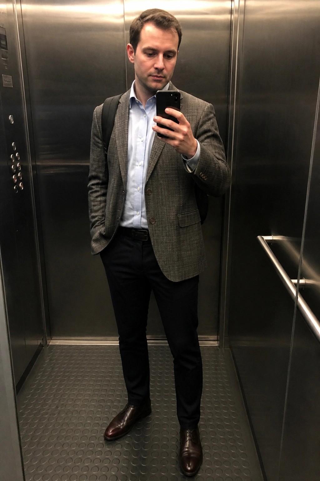 Man in gray plaid blazer over light blue button down shirt, dark navy pants, brown leather dress shoes, and black backpack taking a mirror selfie in an elevator.