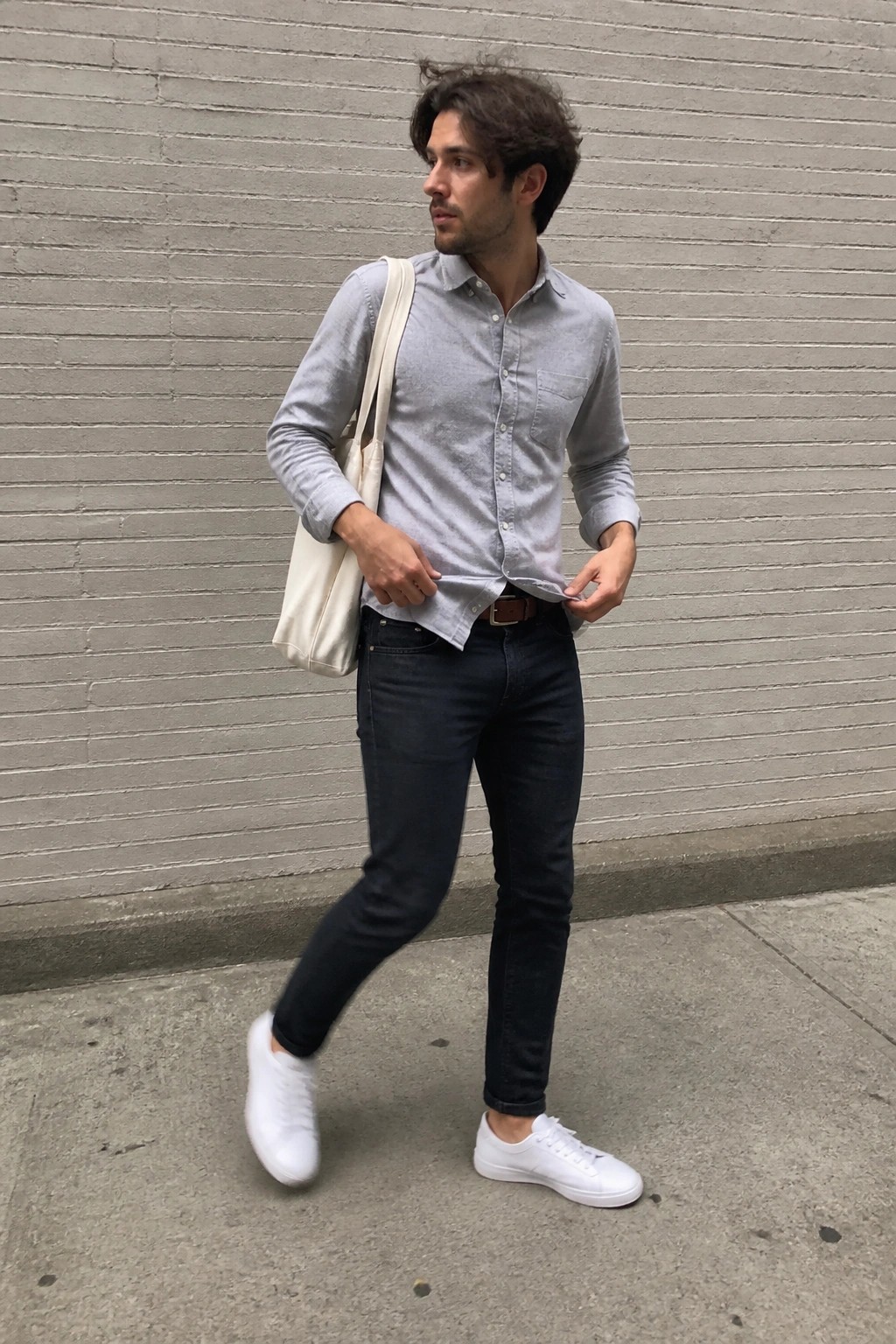 Man in gray shirt and jeans with tote bag against brick wall