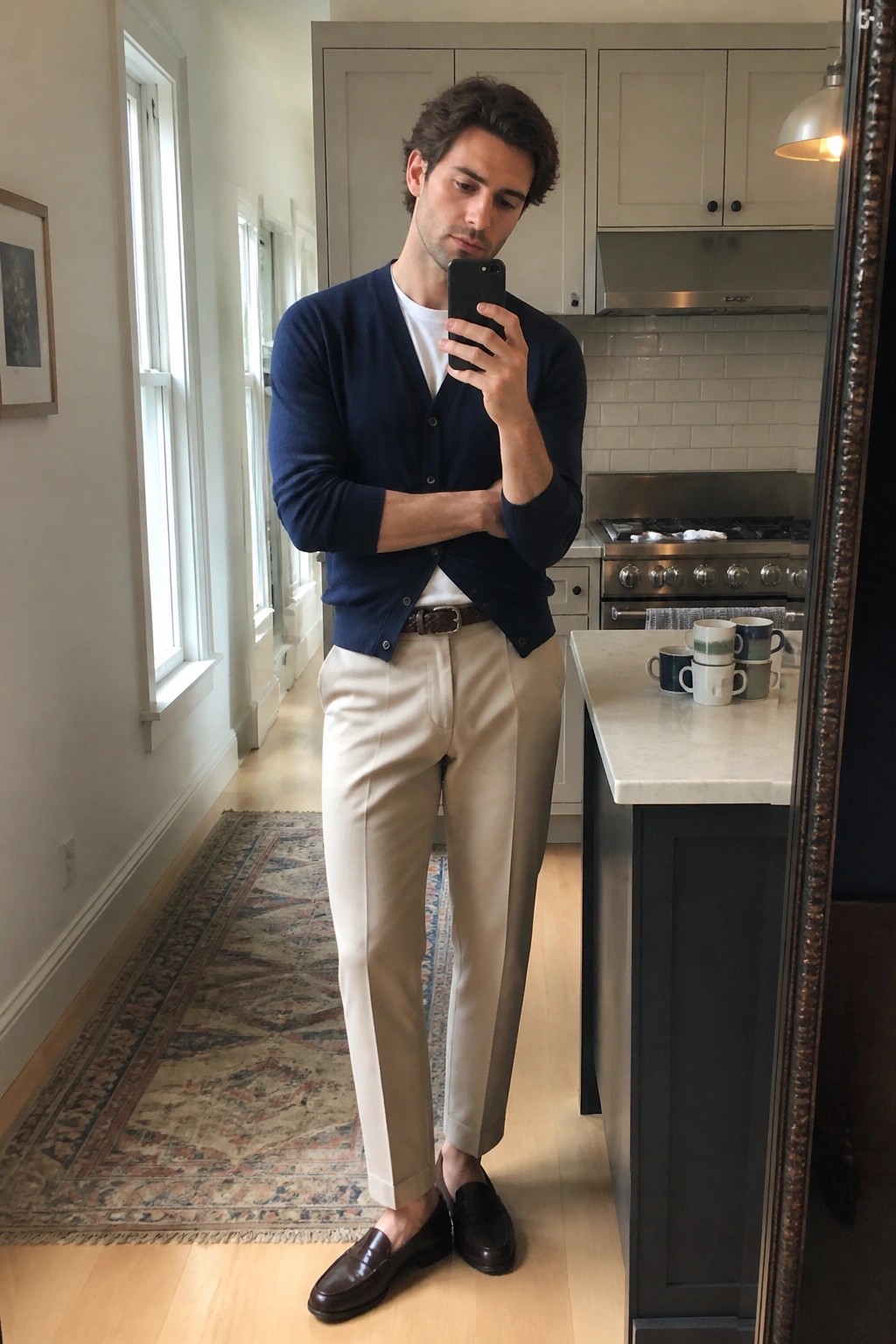 Man in navy cardigan and beige trousers taking mirror selfie in kitchen