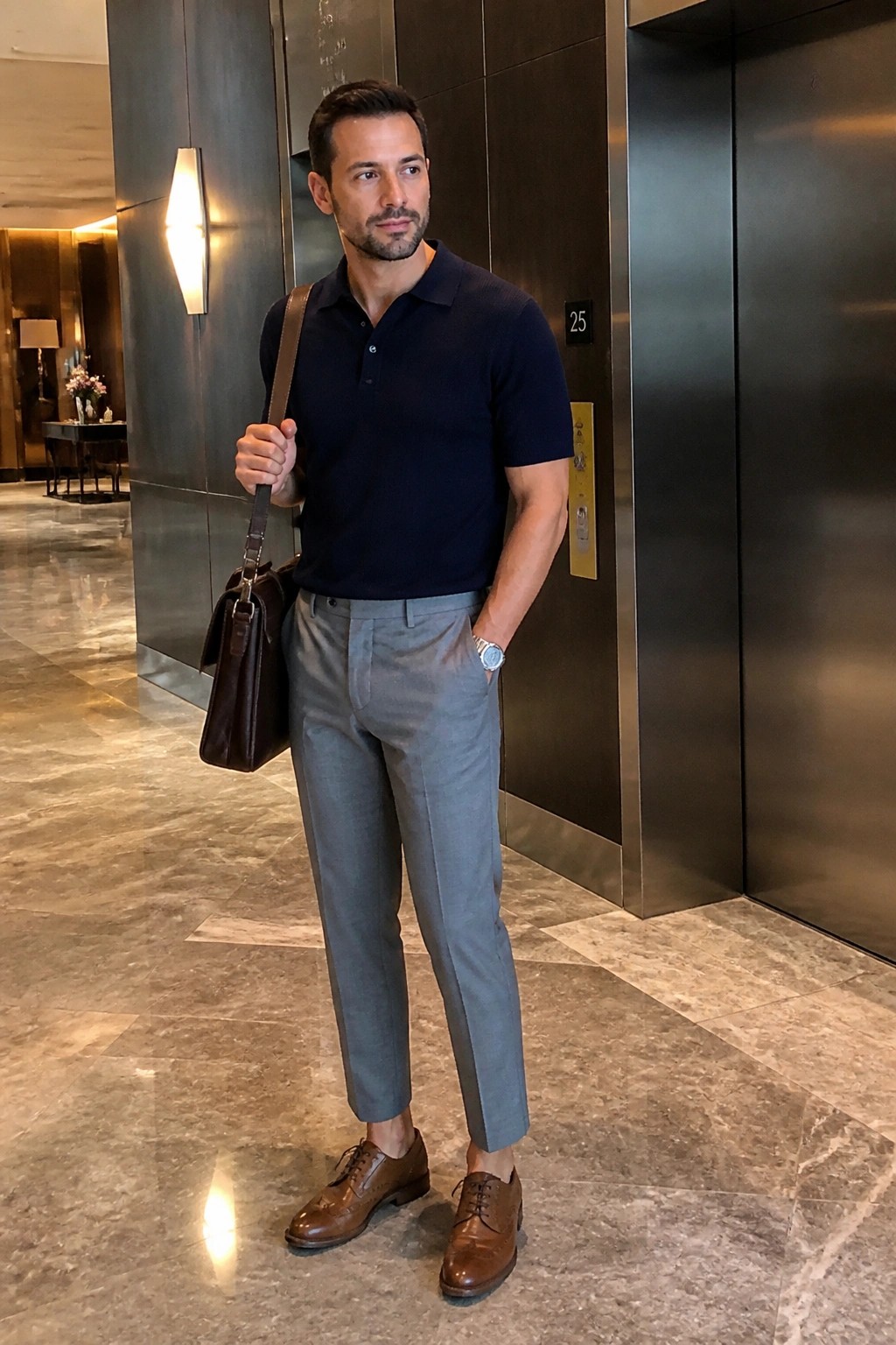 Man in navy polo shirt and gray trousers holding brown leather bag in lobby
