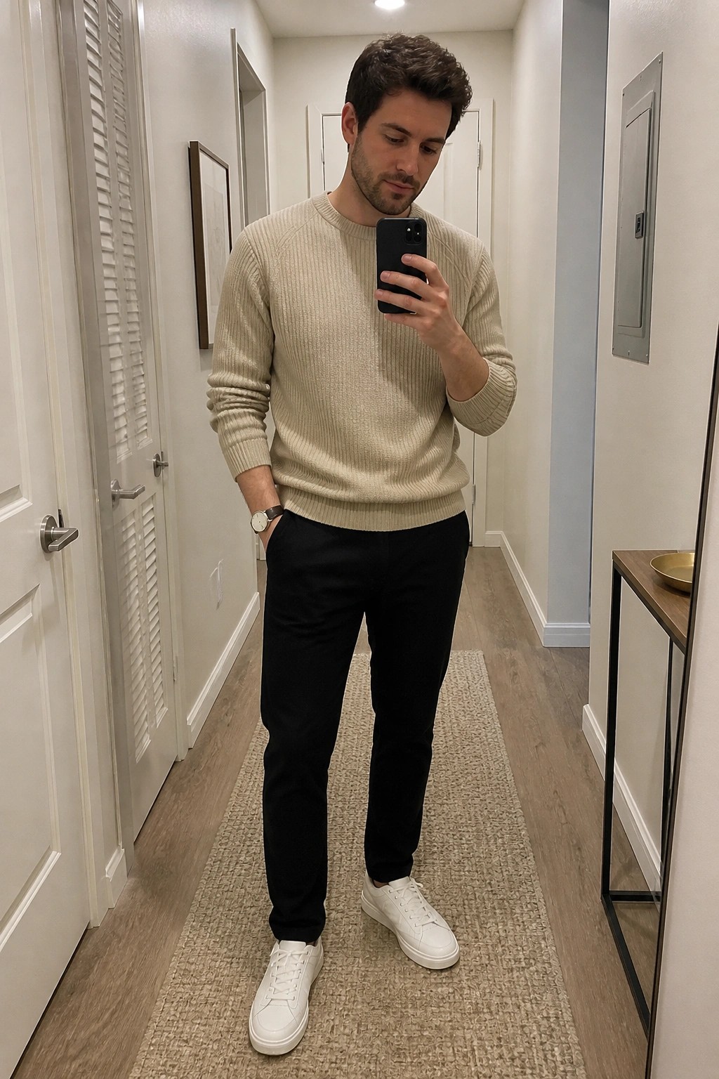 Man in beige sweater and black pants taking mirror selfie in hallway
